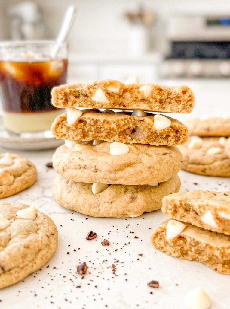 Stack of four light brown coffee cookies with white chocolate chips, two broken in half on top showing soft interior texture. Glass of Vietnamese iced coffee with spoon visible in background, coffee grounds scattered on white surface.