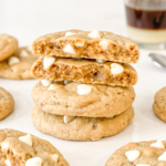 Stack of four coffee-flavored cookies with white chocolate chips, showing the soft, airy texture inside. A glass of layered Vietnamese coffee sits blurred in the background.