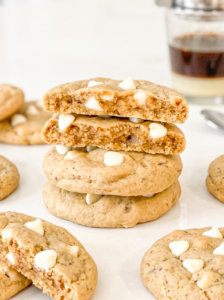 Stack of four coffee-flavored cookies with white chocolate chips, showing the soft, airy texture inside. A glass of layered Vietnamese coffee sits blurred in the background.