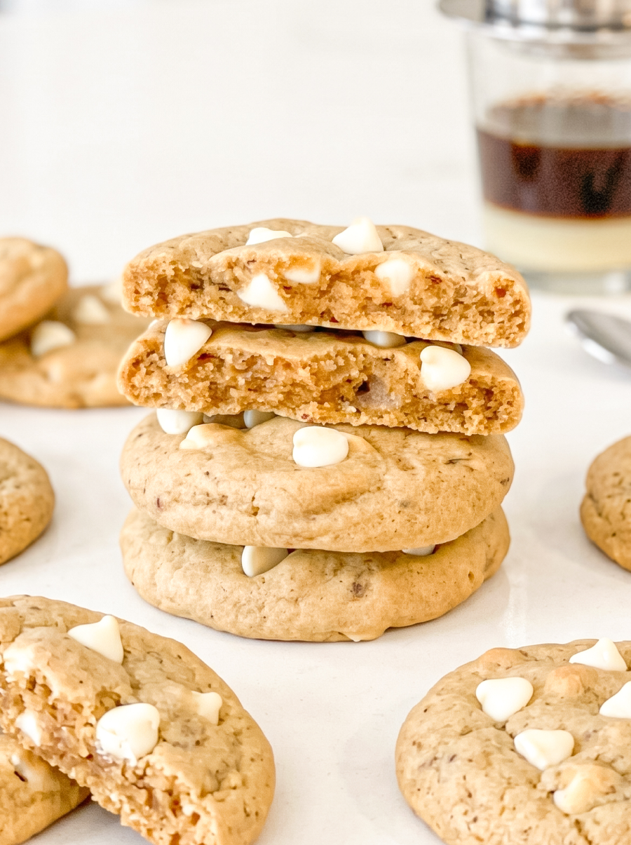 Stack of four coffee-flavored cookies with white chocolate chips, showing the soft, airy texture inside. A glass of layered Vietnamese coffee sits blurred in the background.
