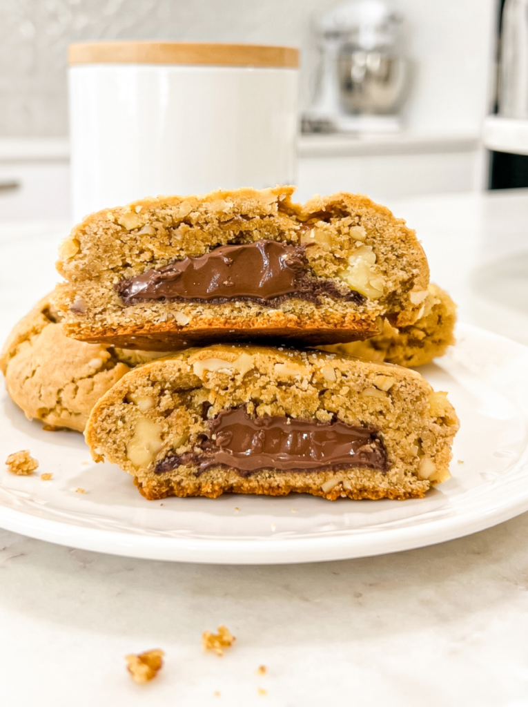 Three walnut cookies stacked on a white plate, two broken in half to reveal gooey Nutella filling inside. Visible walnut pieces dot the golden-brown cookie exterior.