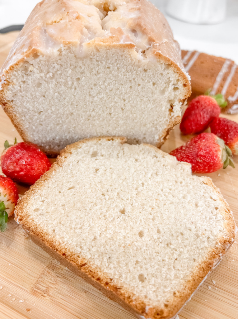Sliced strawberry milk pound cake with glossy white glaze on a wooden cutting board, showing tender white crumb texture. Fresh strawberries garnish the sides, and the glazed loaf stands behind the front slice.