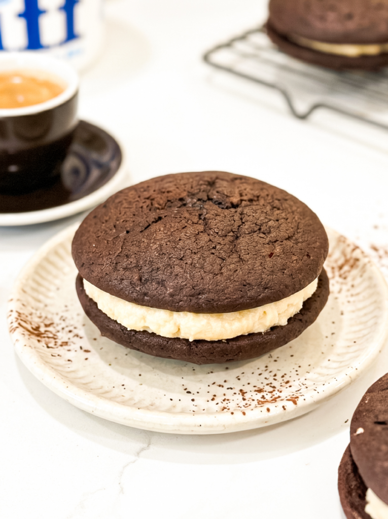 Chocolate whoopie pie with creamy mascarpone filling on a white speckled plate, dusted with cocoa powder, alongside an espresso cup on a marble surface.