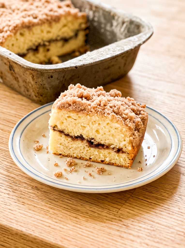 Slice of coffee cake with cinnamon streusel topping and swirl filling on a white plate with blue rim. Rustic loaf pan with remaining cake visible in background on wooden surface.