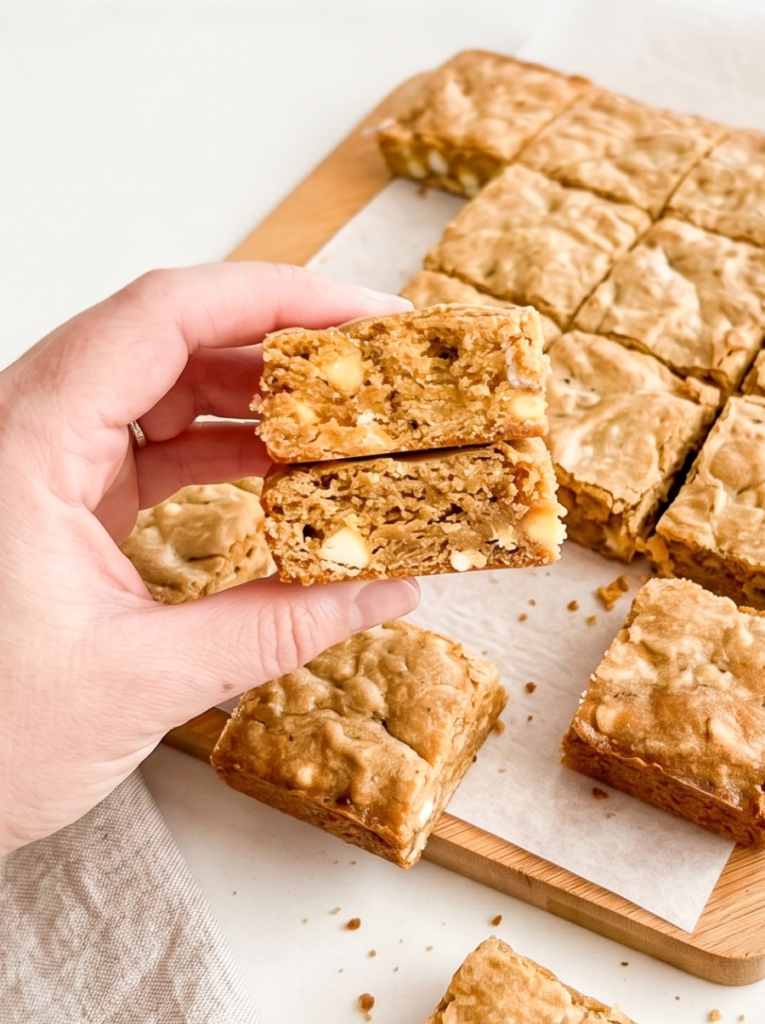 Hand holding two stacked Vietnamese coffee cookie bars broken in half, revealing a golden brown interior studded with white chocolate chips. Additional square-cut bars arranged on a wooden board with parchment paper.