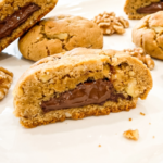 Golden brown walnut cookies broken in half reveal pools of creamy Nutella filling inside, with whole and broken walnuts scattered on a white surface.