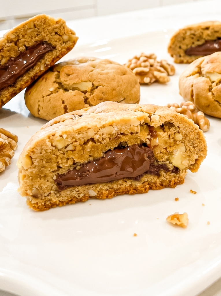 Golden brown walnut cookies broken in half reveal pools of creamy Nutella filling inside, with whole and broken walnuts scattered on a white surface.