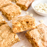 Golden brown coffee-flavored cookies studded with white chocolate chips arranged on parchment paper, with one cookie propped up to show the tender, crumbly interior texture and a small white bowl of white chocolate chips in the background.