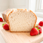 Sliced strawberry milk pound cake on a wooden cutting board showing the tender, fine pink crumb with a sweet glaze on top, surrounded by fresh strawberries against a soft neutral background.