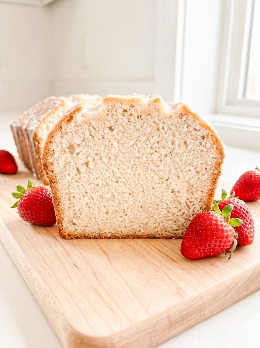 Sliced strawberry milk pound cake on a wooden cutting board showing the tender, fine pink crumb with a sweet glaze on top, surrounded by fresh strawberries against a soft neutral background.