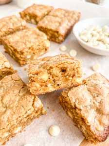 Golden brown coffee-flavored cookies studded with white chocolate chips arranged on parchment paper, with one cookie propped up to show the tender, crumbly interior texture and a small white bowl of white chocolate chips in the background.
