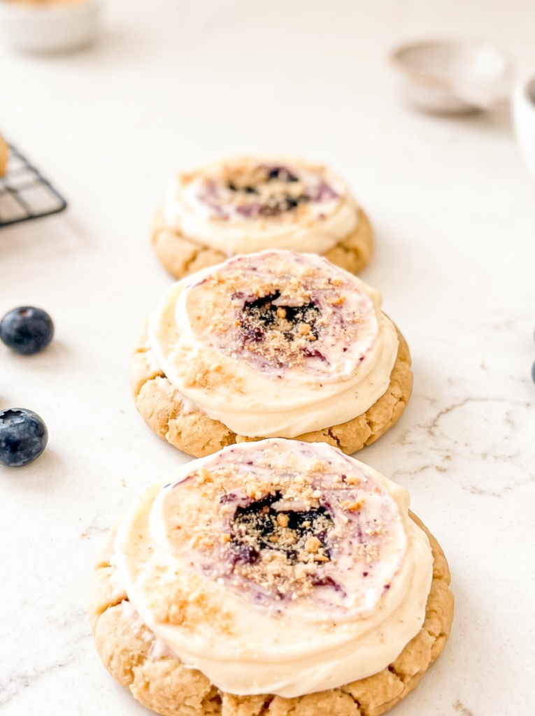 Three soft blueberry cheesecake cookies topped with cream cheese frosting and cookie crumble, arranged in a diagonal line on a white marble surface with fresh blueberries scattered nearby.