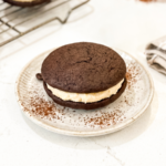 Chocolate whoopie pie with cream filling on a speckled ceramic plate dusted with cocoa powder, photographed from above with a cooling rack and additional whoopie pie in the background.