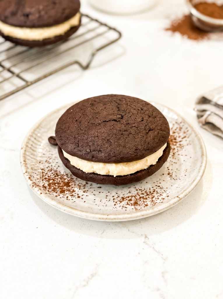 Chocolate whoopie pie with cream filling on a speckled ceramic plate dusted with cocoa powder, photographed from above with a cooling rack and additional whoopie pie in the background.