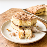 Square slice of coffee cake with thick cinnamon streusel topping on a speckled ceramic plate with a fork, showing moist yellow cake interior with cinnamon swirl layer. Additional cake slices visible on baking sheet in background on rustic wood surface.