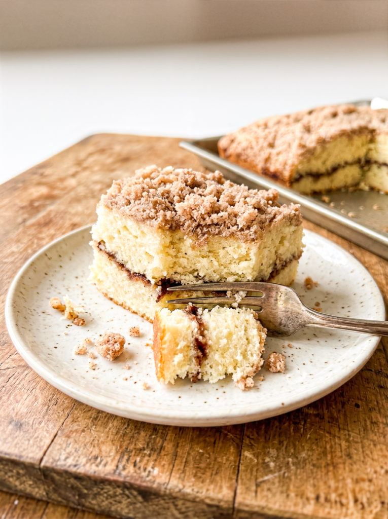 Square slice of coffee cake with thick cinnamon streusel topping on a speckled ceramic plate with a fork, showing moist yellow cake interior with cinnamon swirl layer. Additional cake slices visible on baking sheet in background on rustic wood surface.