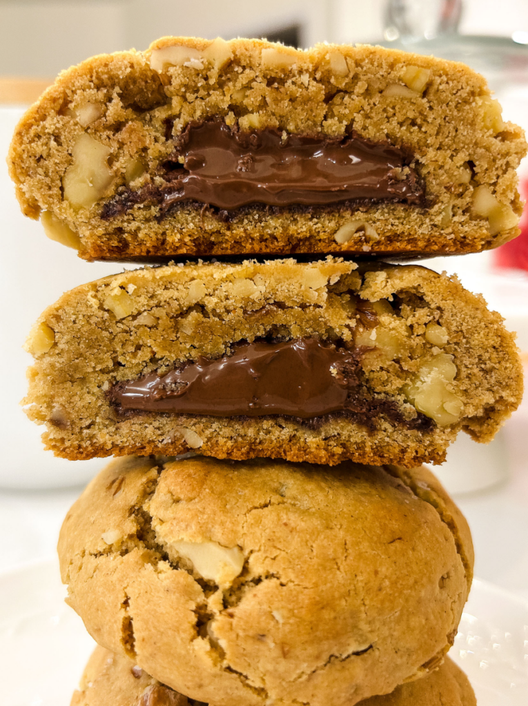 Stack of three walnut cookies with two cut in half to reveal a thick layer of creamy chocolate-hazelnut filling inside, showing the soft golden brown cookie texture studded with chopped walnuts.