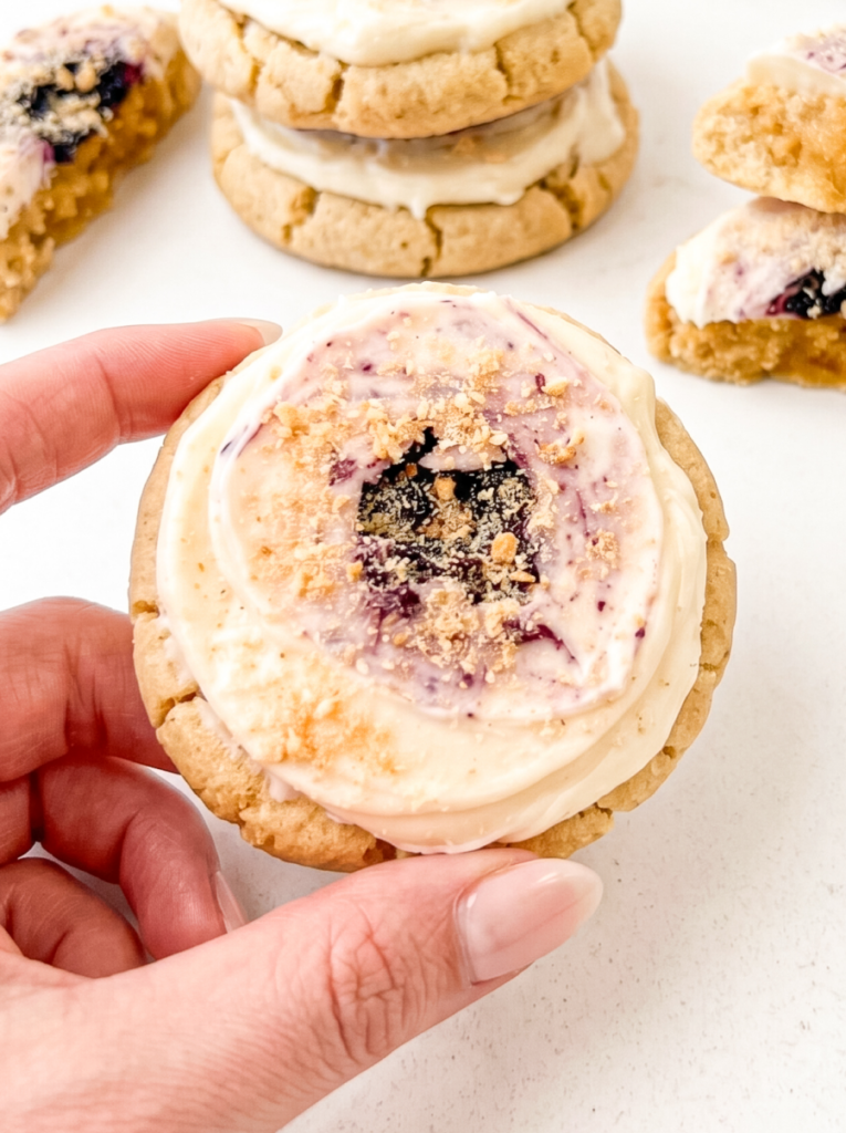 Hand holding a soft sugar cookie topped with swirled cream cheese frosting, purple blueberry jam, and graham cracker crumbs, with more cookies visible in the background on a white marble surface