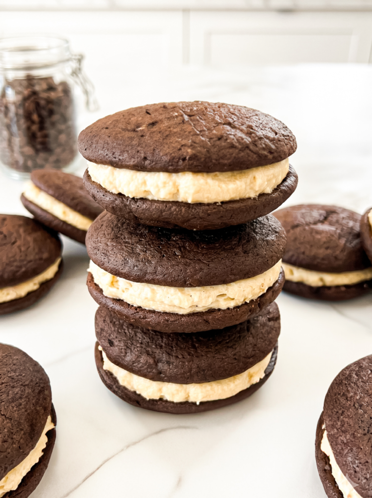 Three tiramisu whoopie pies stacked on a white marble surface, with soft chocolate cookies sandwiching creamy mascarpone filling. Additional whoopie pies are arranged around the stack, and a glass jar of coffee beans sits in the background.