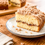 Slice of layered coffee cake on a ceramic plate showing moist yellow cake with cinnamon swirl filling and thick crumb topping, with whole cake and serving board visible in background on wooden table