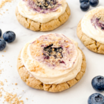 Soft golden cookies topped with cream cheese frosting swirled with blueberry jam and cookie crumbs, arranged on a black wire cooling rack with fresh blueberries scattered around.