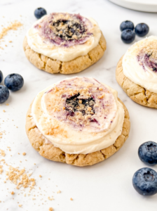 Soft golden cookies topped with cream cheese frosting swirled with blueberry jam and cookie crumbs, arranged on a black wire cooling rack with fresh blueberries scattered around.