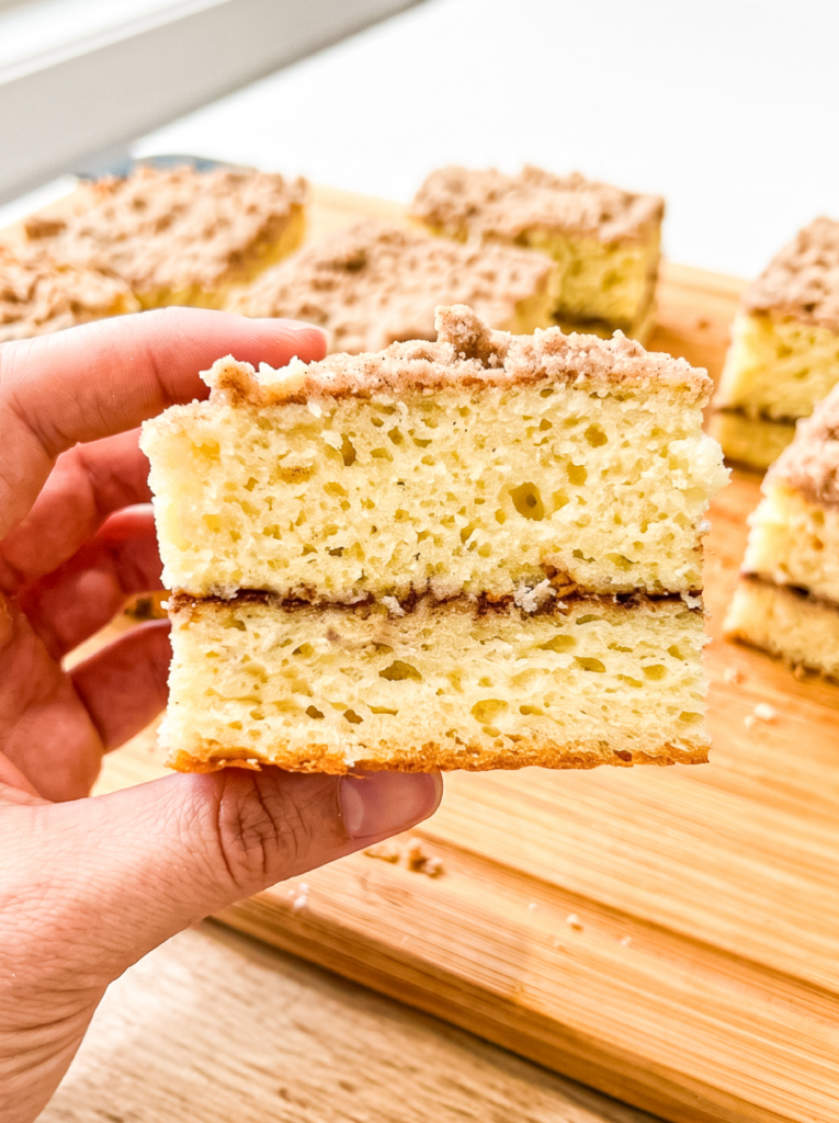 Hand holding two stacked squares of coffee cake with cinnamon streusel topping, showing the tender, moist yellow crumb. More cake squares rest on a wooden cutting board in the background.