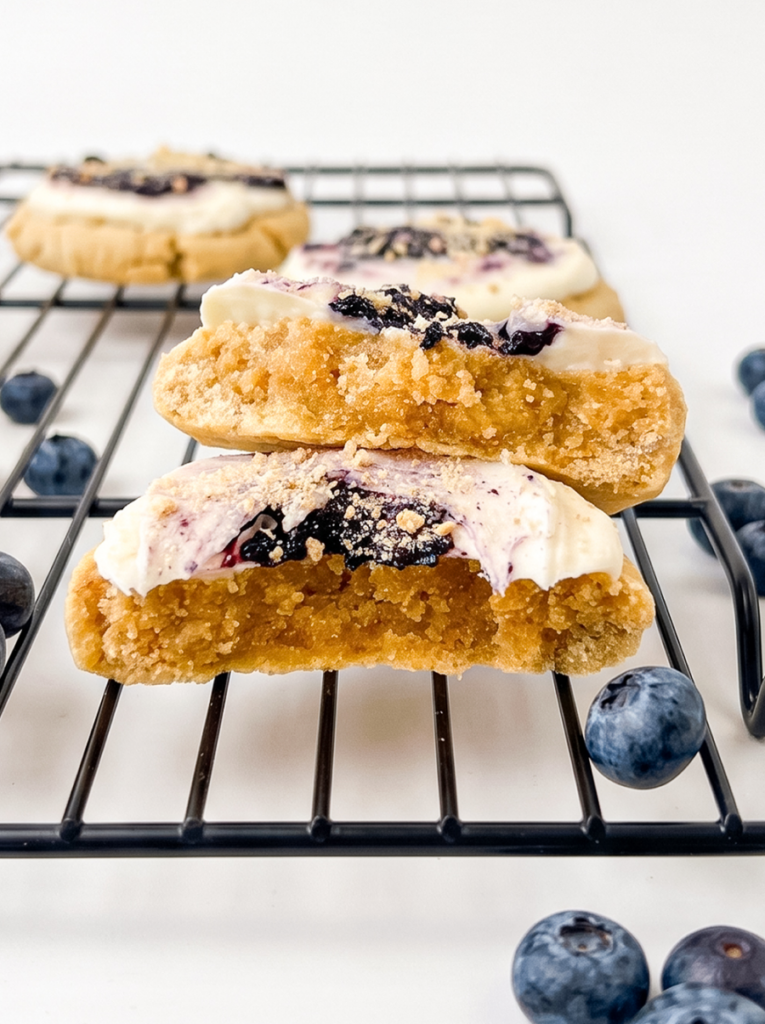 Two thick golden cookies stacked on a black wire cooling rack, topped with cream cheese frosting and dried blueberries, with fresh blueberries scattered around on a white surface