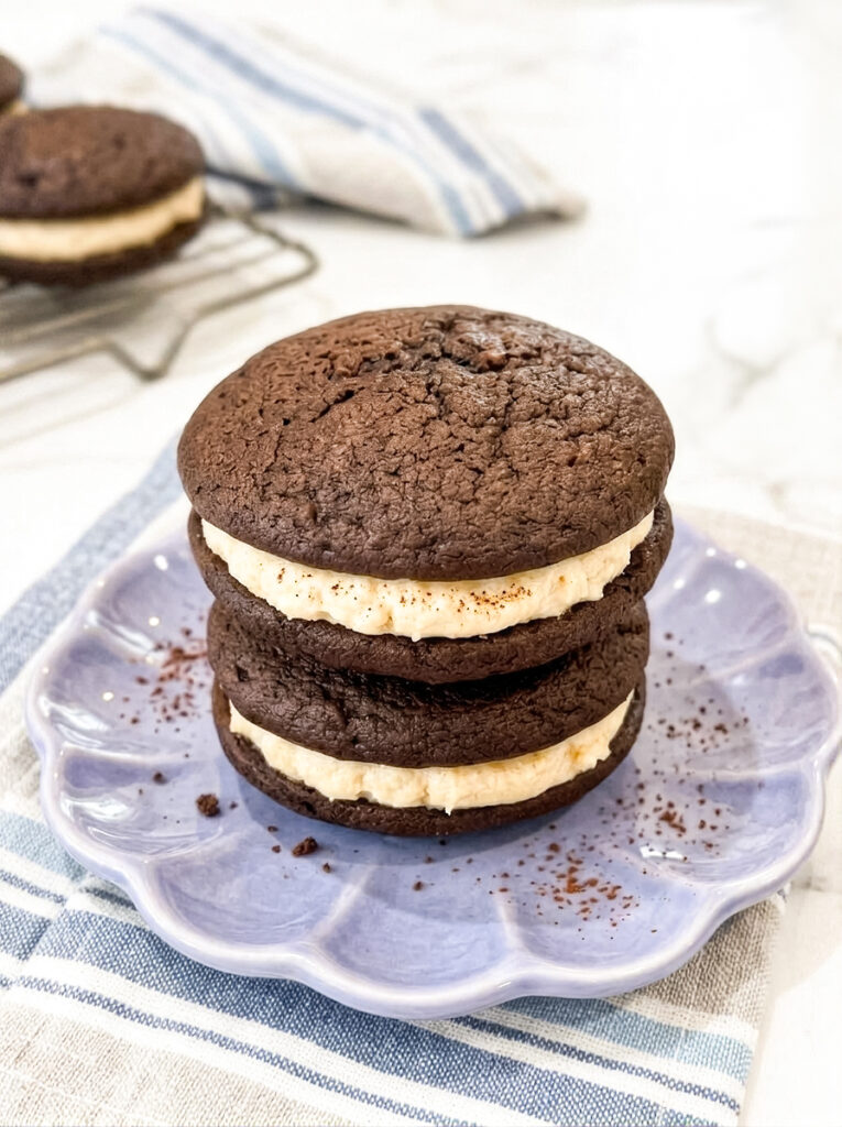 A stack of two chocolate whoopie pies filled with cream sits on a purple scalloped plate with loose cocoa powder on a striped napkin.