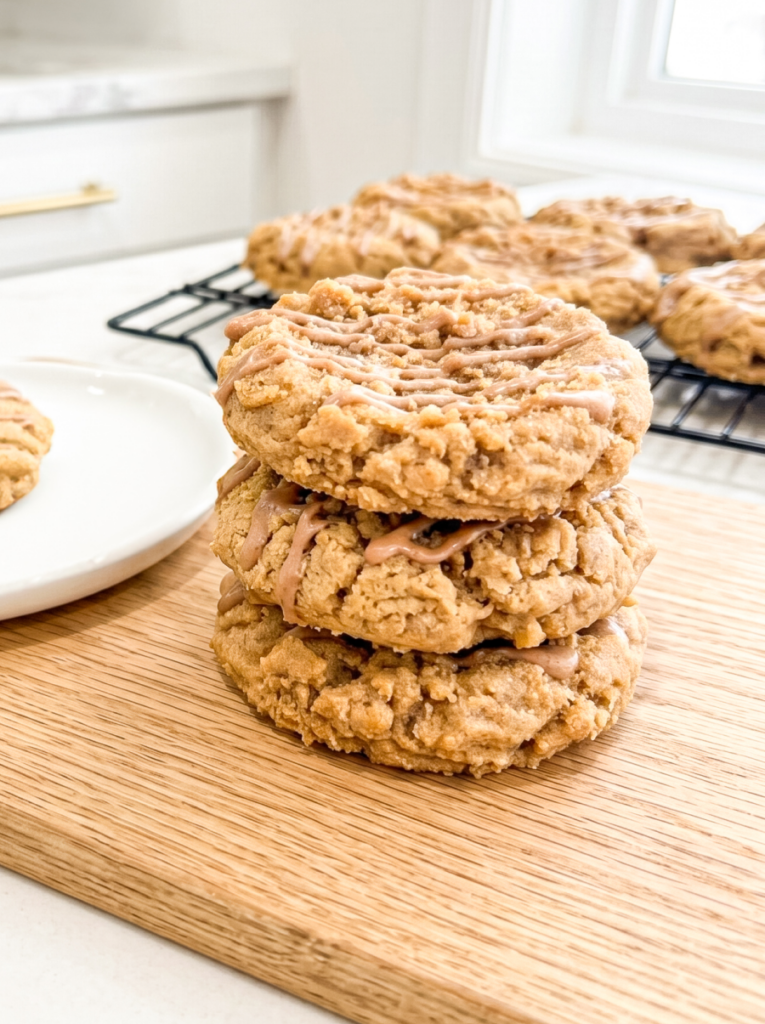 Stack of three thick coffee cake cookies with crumbly streusel topping and vanilla glaze drizzled on top, displayed on a bamboo board with more cookies cooling on a black wire rack in the background