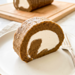Slice of brown hojicha sponge cake rolled with white cream filling, plated on white dish with gold rim and fork. Full cake roll visible on wooden board in background.