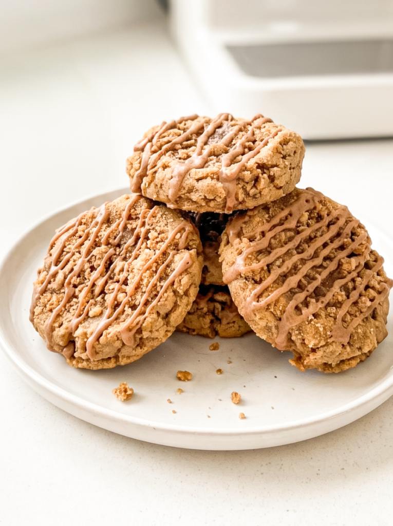 Stack of five golden-brown cookies with crumbly streusel topping and cinnamon glaze drizzled across them, arranged on a cream-colored plate with a blurred white mug in the background