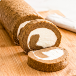 Sliced hojicha cake roll on a wooden cutting board showing golden-brown roasted tea sponge cake spiraled around creamy white filling, with a white kitchen towel in the background.