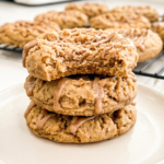 Stack of three thick coffee cake cookies on a white plate showing crumbly streusel topping and cinnamon drizzle, with a bite taken from the top cookie revealing soft interior. More cookies on a cooling rack in the background.