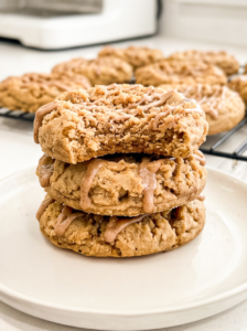 Stack of three thick coffee cake cookies on a white plate showing crumbly streusel topping and cinnamon drizzle, with a bite taken from the top cookie revealing soft interior. More cookies on a cooling rack in the background.