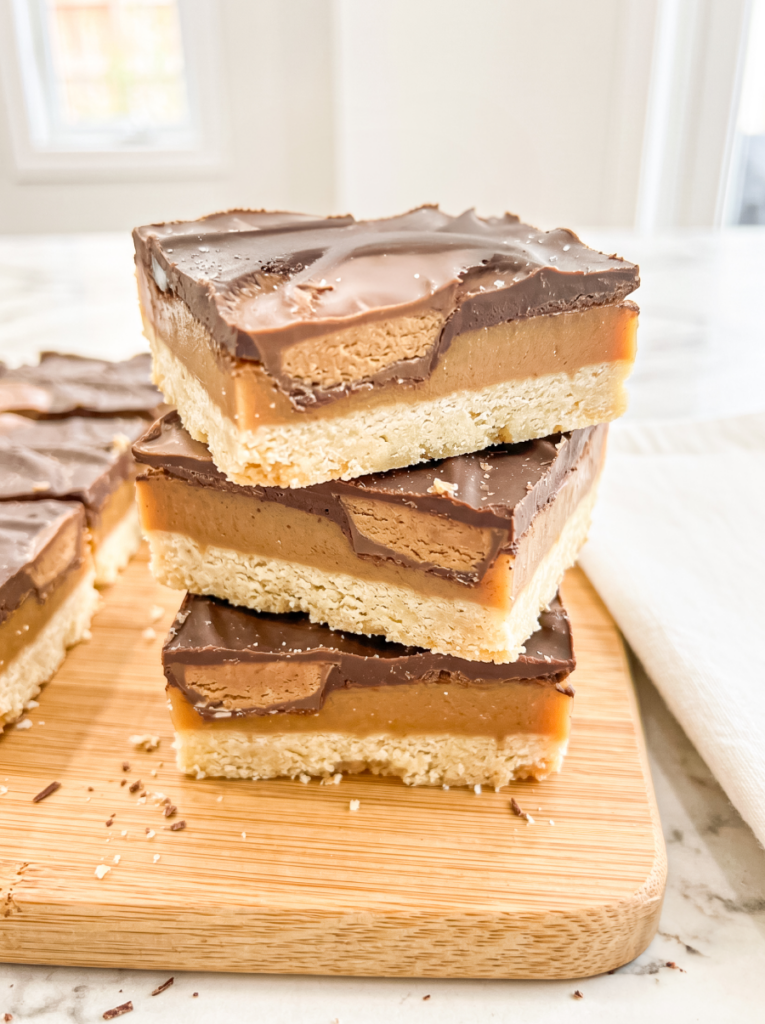 Three layered dessert bars stacked on a bamboo cutting board showing a shortbread base, caramel center, peanut butter layer, and chocolate topping, with additional bars and chocolate pieces in the background.