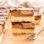 Two stacked Reese millionaire bars on a wooden board showing distinct layers: shortbread base, caramel center, peanut butter filling, and chocolate top. A silver fork rests beside the bars with scattered crumbs visible.