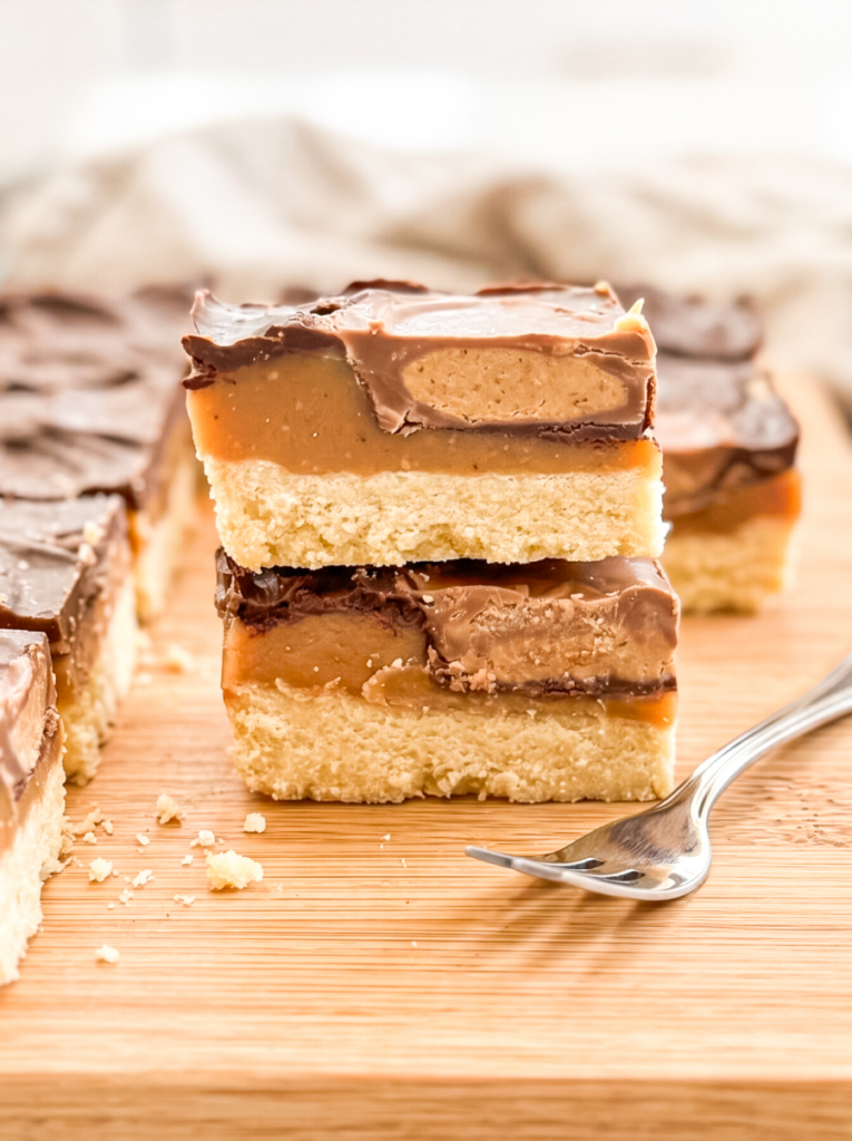 Two stacked Reese millionaire bars on a wooden board showing distinct layers: shortbread base, caramel center, peanut butter filling, and chocolate top. A silver fork rests beside the bars with scattered crumbs visible.