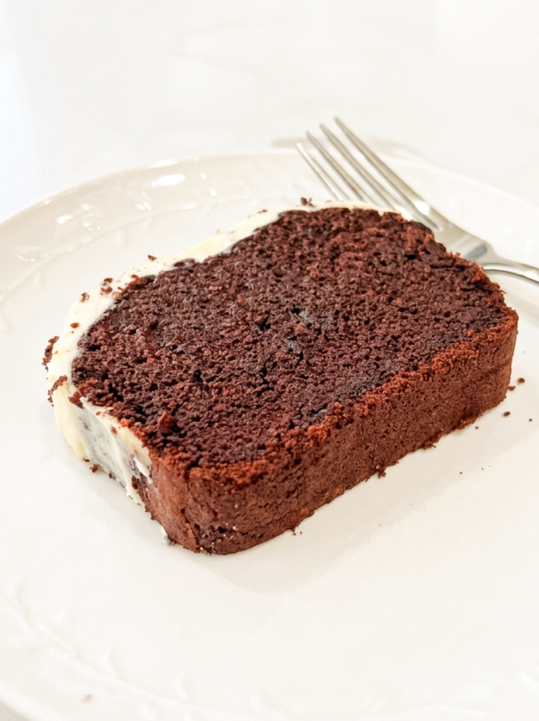 Single slice of red velvet loaf cake on a white plate with a silver fork, showing the deep reddish-brown tender crumb and moist texture.