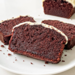 Sliced red velvet loaf cake with cream cheese frosting on a white plate, showing the moist reddish-brown crumb texture. Three slices arranged in foreground with the remaining loaf visible behind.