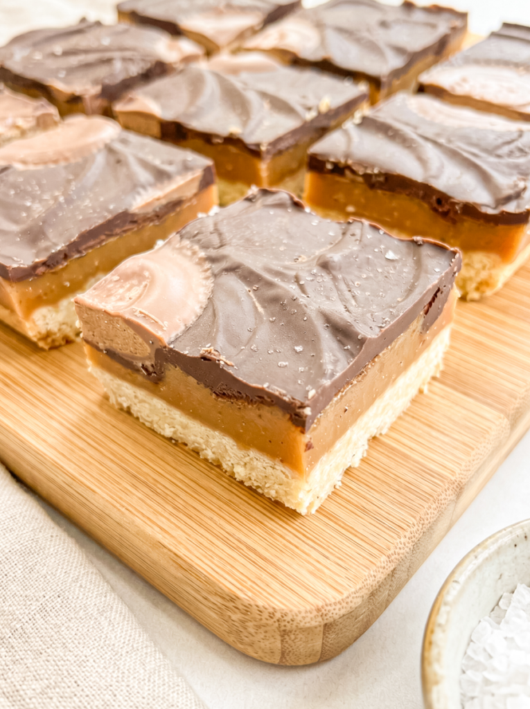 Layered dessert bars on a wooden cutting board showing a shortbread base, golden caramel center, and smooth milk chocolate top with swirled finish. One bar is positioned in front with additional bars arranged behind.