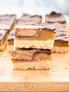 Two layered dessert bars stacked on a wooden board showing a shortbread base, golden caramel layer, peanut butter filling, and milk chocolate top, with additional bars blurred in the background.
