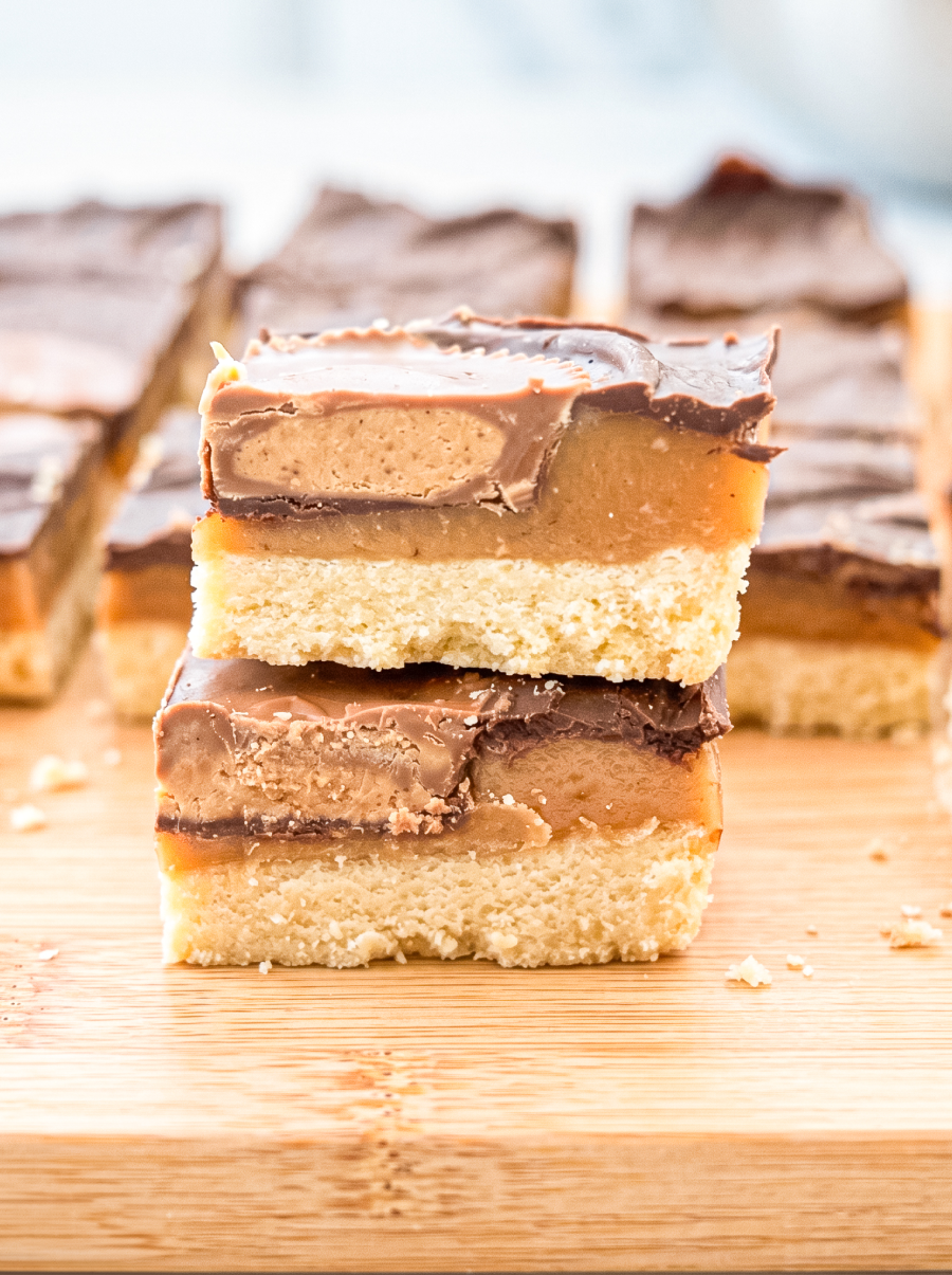 Two layered dessert bars stacked on a wooden board showing a shortbread base, golden caramel layer, peanut butter filling, and milk chocolate top, with additional bars blurred in the background.