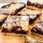 Stack of blueberry crumble bars on a wooden cutting board showing golden crust, dark purple blueberry filling, and buttery crumble topping dusted with powdered sugar. A vintage silver spoon rests in the foreground.