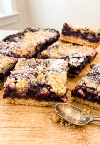 Stack of blueberry crumble bars on a wooden cutting board showing golden crust, dark purple blueberry filling, and buttery crumble topping dusted with powdered sugar. A vintage silver spoon rests in the foreground.