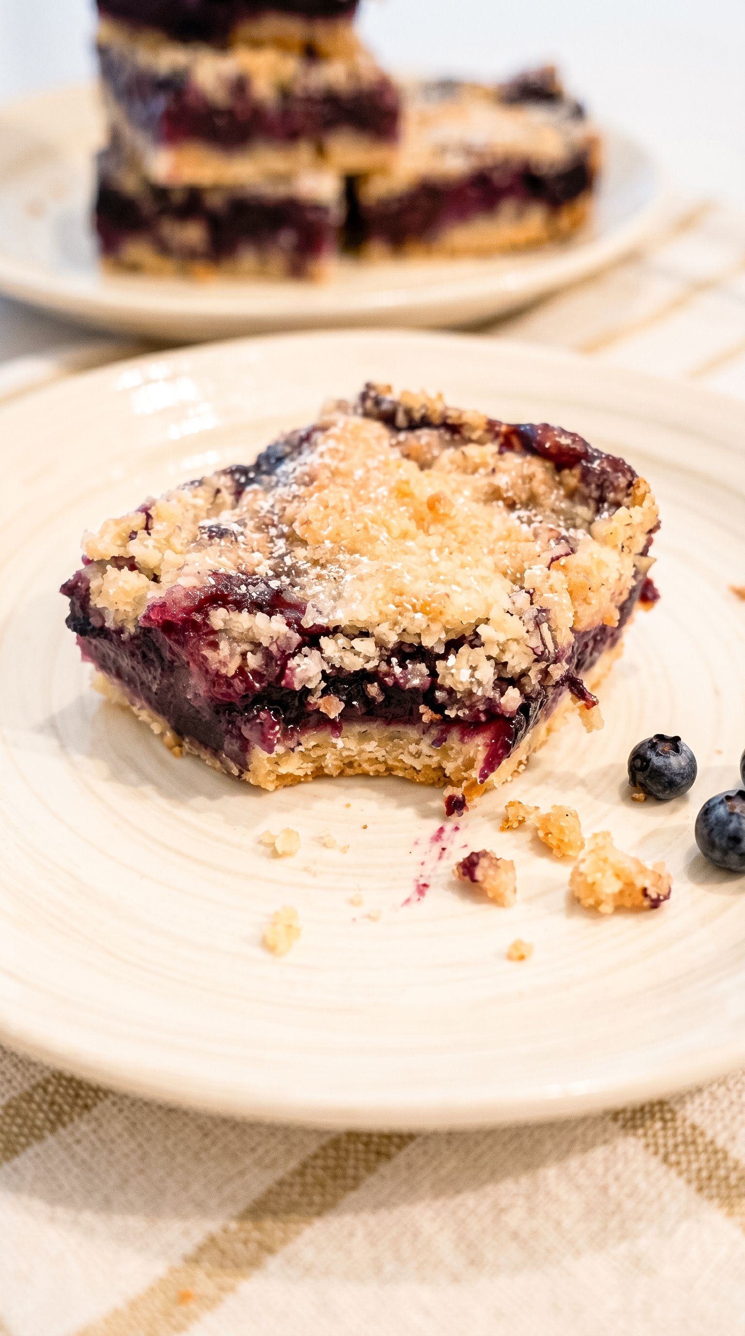 A single blueberry crumble bar on a white plate showing golden buttery crust, thick dark purple blueberry filling, and crumbly topping with scattered crumbs and fresh blueberries beside it. More bars stacked on a plate in the background.