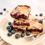 Three stacked blueberry crumble bars on a wooden plate with a fork, showing golden crumbly topping and deep purple blueberry filling, surrounded by fresh blueberries.
