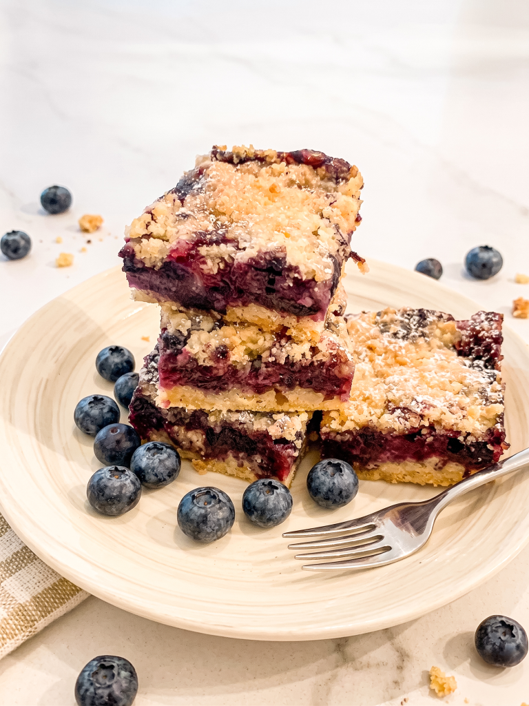 Three stacked blueberry crumble bars on a wooden plate with a fork, showing golden crumbly topping and deep purple blueberry filling, surrounded by fresh blueberries.