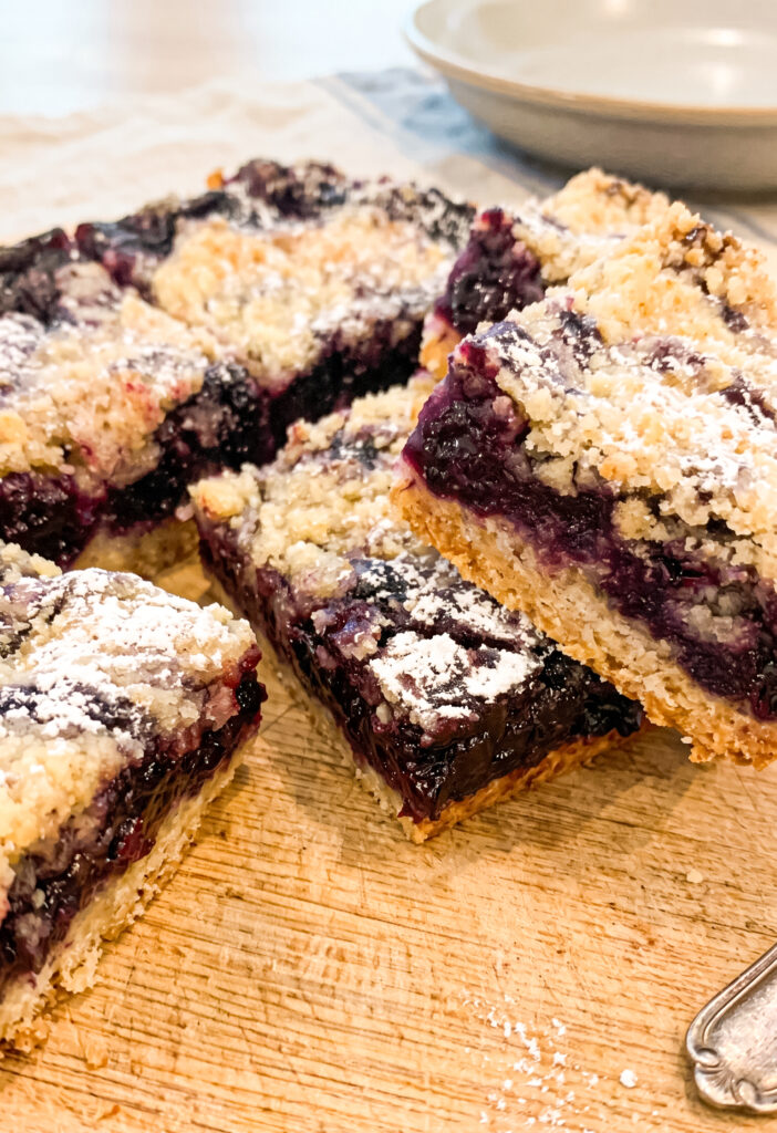 Four golden crumble bars with deep purple blueberry filling arranged on a wooden cutting board, dusted with powdered sugar and photographed from above with cream-colored plates in the background.