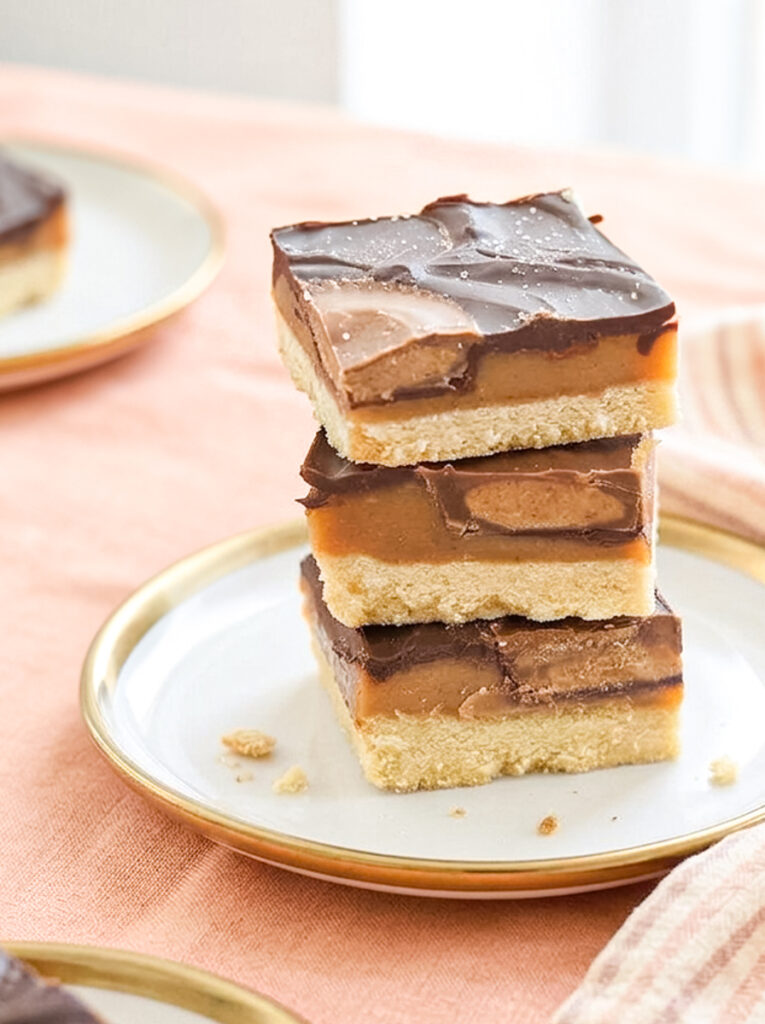 Three chocolate caramel shortbread bars with peanut butter cups stacked on a small white plate with a gold rim.